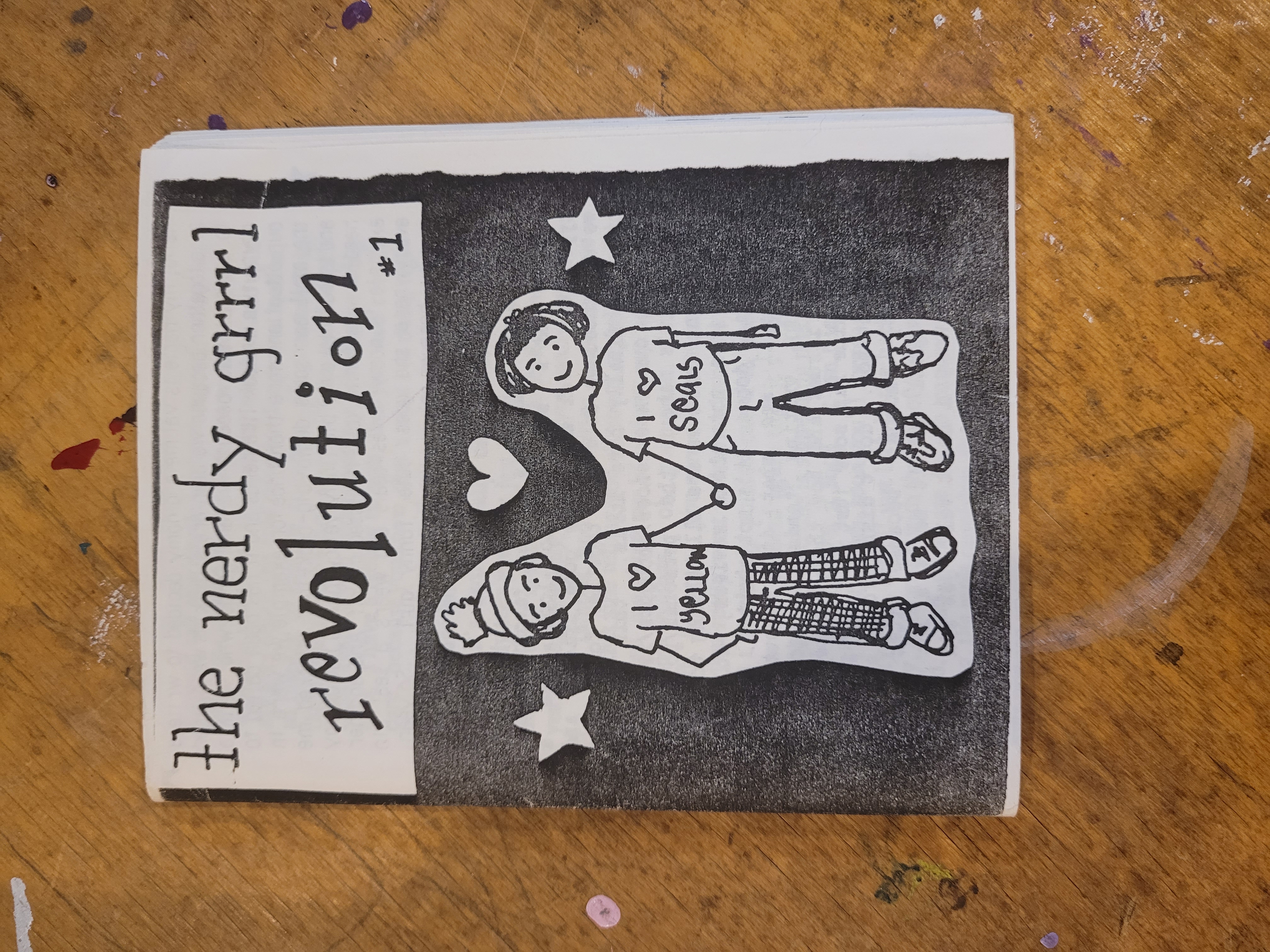 cover of black and white photocopied zine whihc reads in handwritten font 'the nerdy grrrl revolution' with a drawing of two people holding hands. One with a shirt that says 'I love yeallow' and one with a shirt saying 'I love seals'