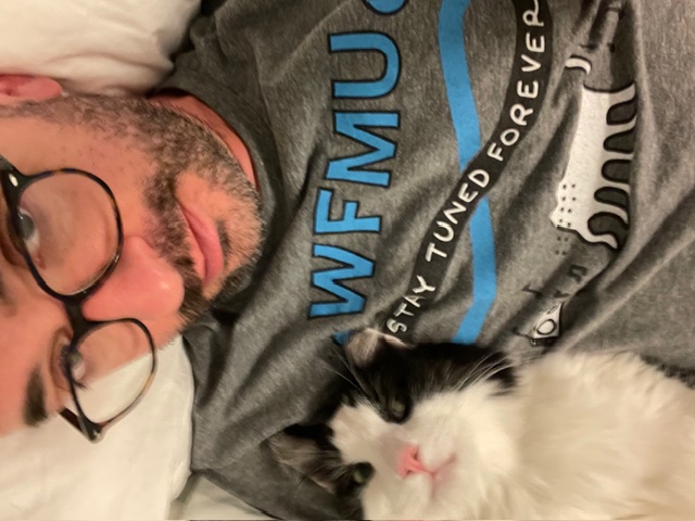 Person with glasses, beard, and grey WFMU t-shirt sitting with a black and white cat